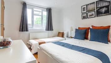 A hotel room with two beds, a white desk, a bowl of fruit, and a window with curtains.