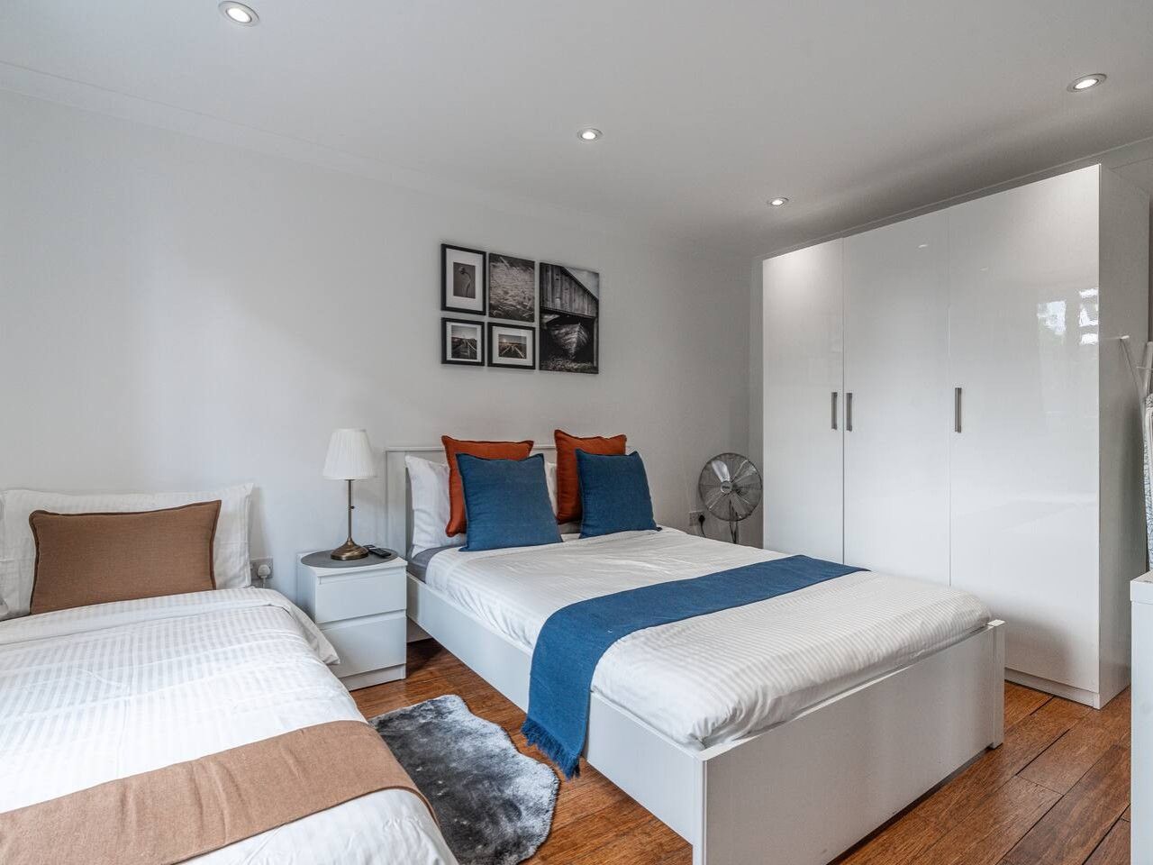 Quarto com duas camas, um guarda-roupa, mesa de cabeceira com abajur e ventoinha na parede.