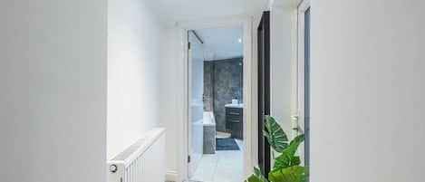A hallway with a radiator, a potted plant, and an open door leading to a bathroom with a shower and a sink.
