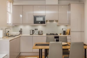 A modern kitchen with a built-in oven, a coffee maker, and a dining area with a table and chairs. - Fitzrovia Apartments | 3BR | Sleeps 12 | Oxford-2 (Greater London)