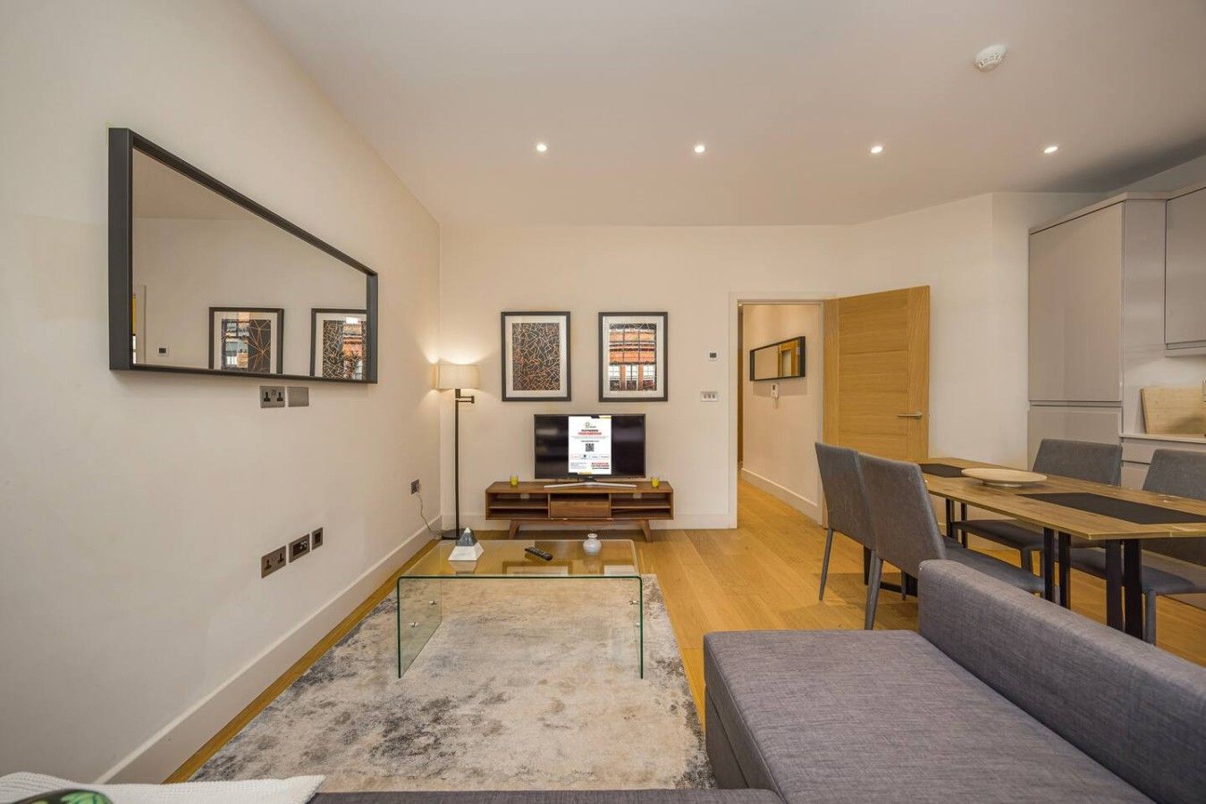 A modern living room with a flat-screen TV, a sofa, a dining table with chairs, and a kitchen area.