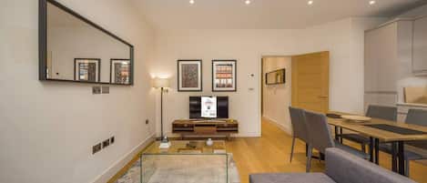 A modern living room with a flat-screen TV, a sofa, a dining table with chairs, and a kitchen area.