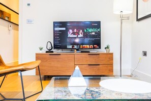 A modern living room with a flat-screen TV on a wooden cabinet, a glass coffee table, and a chair. - Fitzrovia 1BR Retreat | Near Soho & Tube-13 (Greater London)