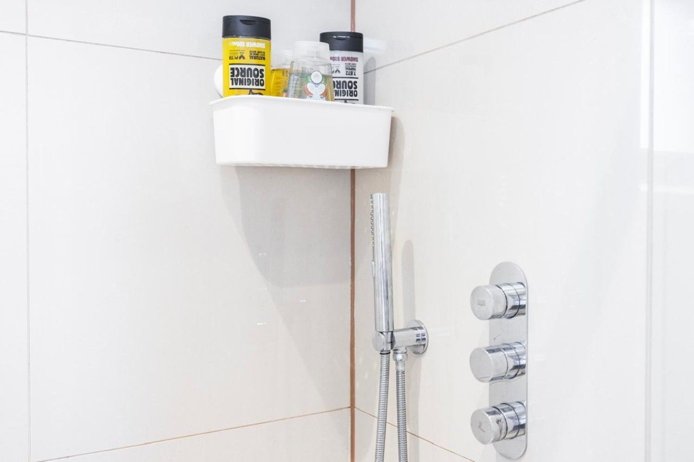 Ein Badezimmer mit weiß gefliester Wand, einer Dusche mit Chromarmaturen und Ablage für Toilettenartikel.