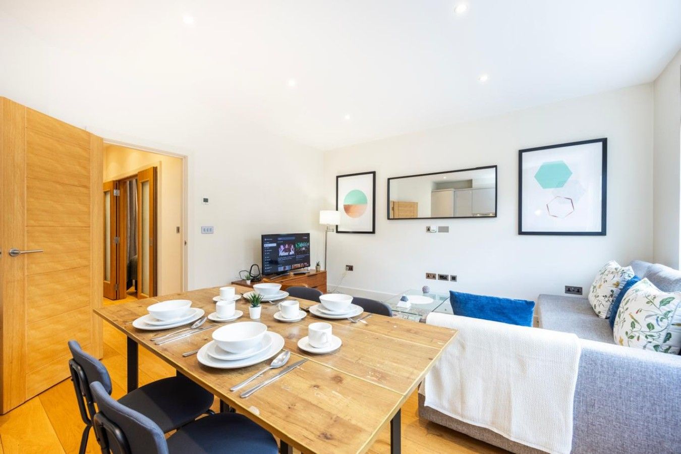 A dining area with a wooden table set for four, a television, and a sofa with a patterned throw.