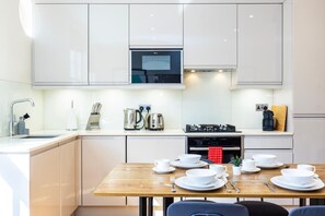 A modern kitchen with white cabinets, a stainless steel stove, and a wooden dining table set with white dishes. - Fitzrovia 1BR Retreat | Near Soho & Tube-13 (Greater London)