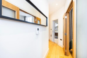 A hallway with a white wall, a wooden floor, a mirror, and a door leading to a bathroom. - Fitzrovia 1BR Retreat | Near Soho & Tube-13 (Greater London)