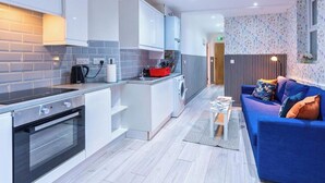 A modern kitchen with a built-in oven and a compact island, a blue sofa with cushions, and a patterned wallpaper on the adjacent wall. - Stylish 3BR | 3 En-Suite Showers | Sleeps 8 (Greater London)