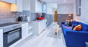 A modern kitchen with a built-in oven and a compact island, a blue sofa with cushions, and a patterned wallpaper on the adjacent wall. - Stylish 3BR | 3 En-Suite Showers | Sleeps 8 (Greater London)