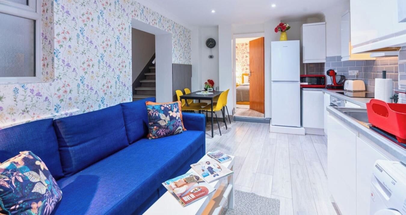 Een knus woongedeelte met een blauwe bank, een eettafel met stoelen, een keuken met een koelkast, en een trap naar een aangrenzende kamer.