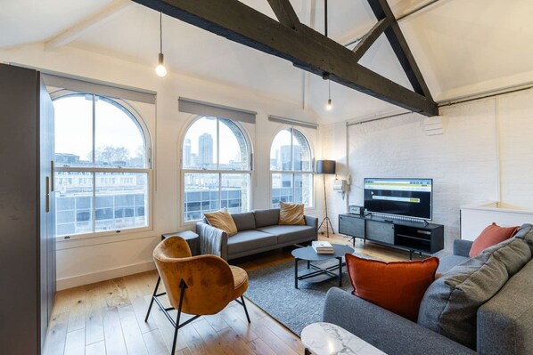 A modern living room with a sofa, armchairs, a coffee table, and a television. Large windows provide views of the cityscape. - Farringdon Loft | Balcony | Sleeps 4 | Zone 1 (Greater London)