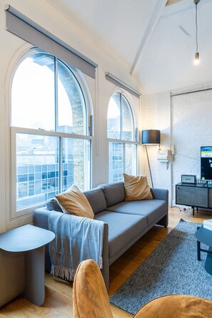 A modern living room with a grey sofa, a small round table, and a large window with a view of the city. - Farringdon Loft | Balcony | Sleeps 4 | Zone 1 (Greater London)