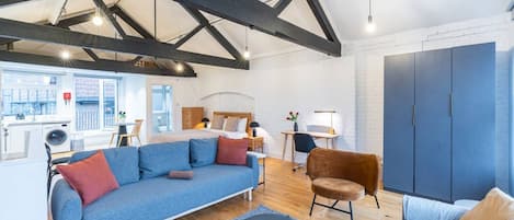 Una sala moderna con un sofá azul, un sillón marrón y un tapete gris. Se observa una mesa de centro de madera y una cama con cabecera blanca al fondo.