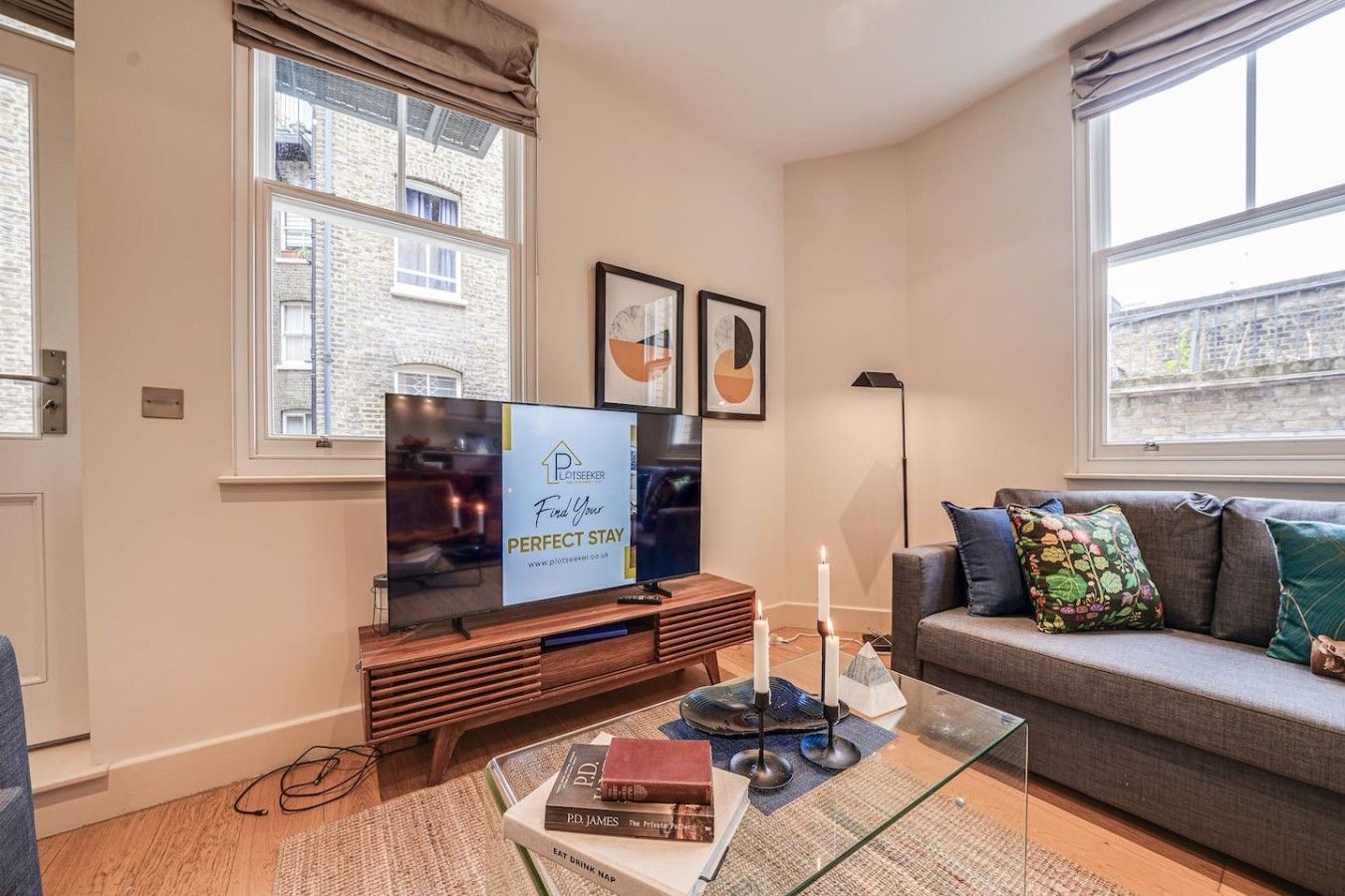 A modern living room with a flat-screen TV, a sofa with cushions, a coffee table with books and candles, and a window with a view of buildings.