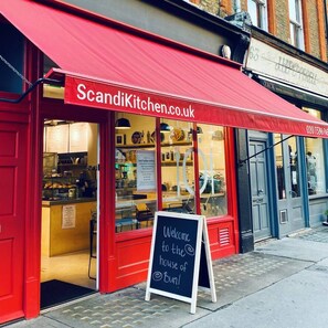 Eine rote Markise mit dem Schriftzug 'ScandiKitchen.co.uk' und ein Schild, das 'Willkommen im Bun-Haus!' verkündet.