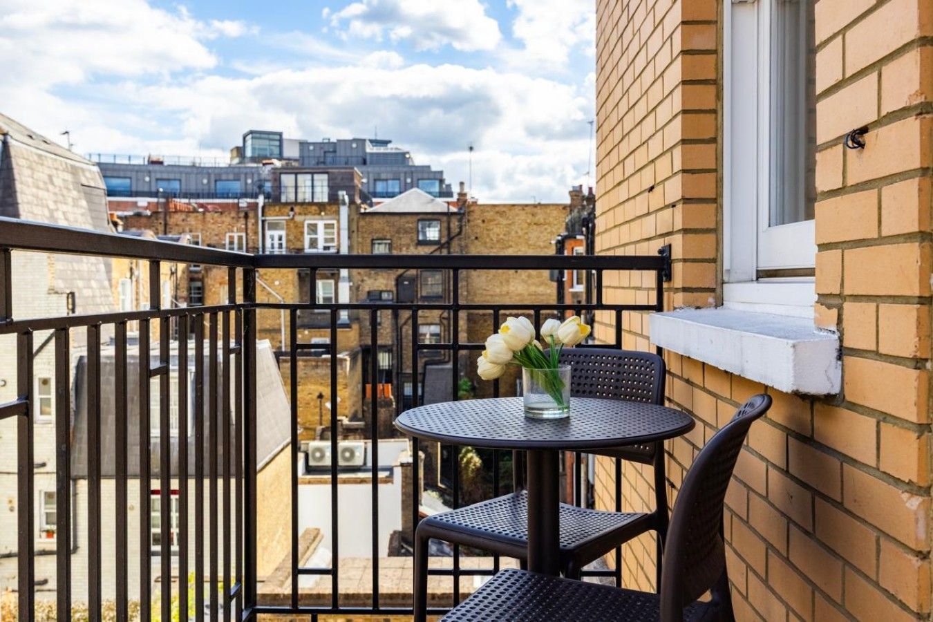 A balcony with a round table and two chairs, a vase with flowers, and a view of buildings in the background.