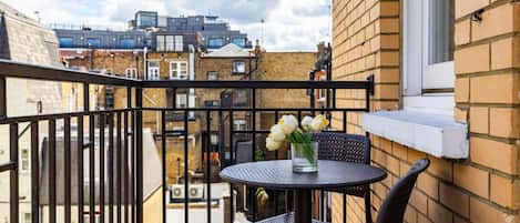 A balcony with a round table and two chairs, a vase with flowers, and a view of buildings in the background.