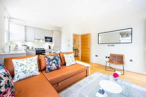 Ein modernes Wohnzimmer mit einer Couch, einem Couchtisch und einem Sessel. Im Hintergrund befindet sich eine Küche mit weißen Schränken und einer Holztür.