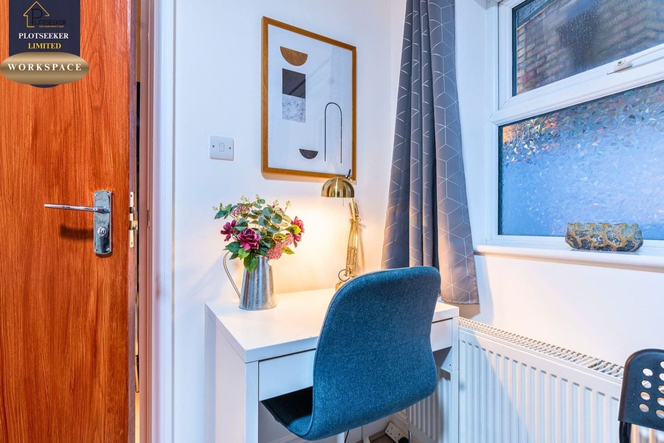 A room with a wooden door, a desk with a chair, a radiator, a window with curtains, and a framed picture on the wall.