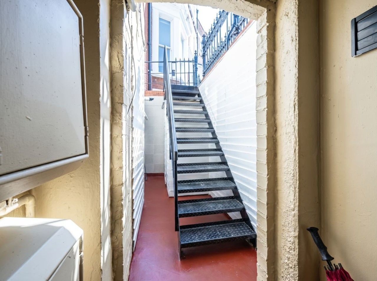 A narrow stairway leading to a landing, flanked by white walls and a red floor, with a black railing and a white appliance on the left.