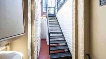 A narrow stairway leading to a landing, flanked by white walls and a red floor, with a black railing and a white appliance on the left.