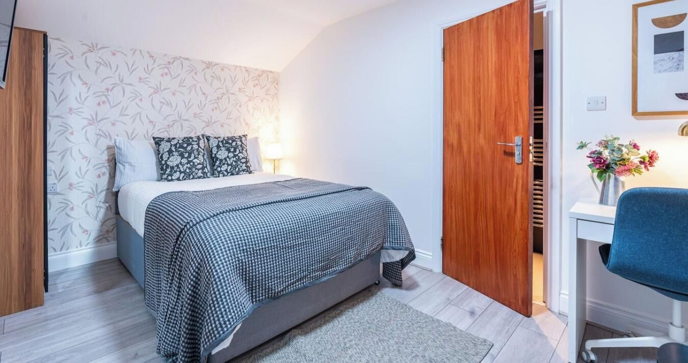 A bedroom with a bed, a bedside table, a chair, a wooden door, and a wall with a floral pattern.