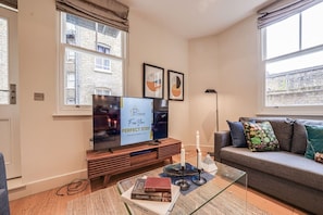 A modern living room with a flat-screen TV, a sofa with cushions, a coffee table with books and candles, and a window with a view of buildings. - Fitzrovia 1BR Retreat | Near Soho & Tube-6 (Greater London)