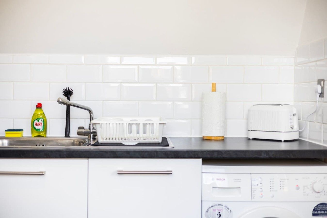 A kitchen with a white tiled backsplash, a Bosch dishwasher, a white toaster, a dish rack, a dish soap bottle, and a dish brush.