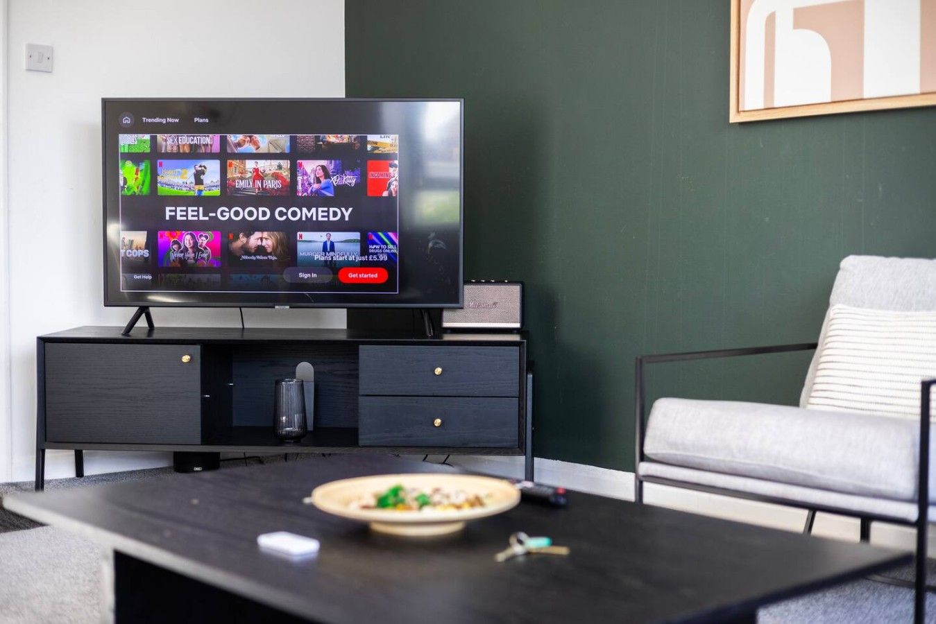 A modern living room with a flat-screen TV displaying a streaming service interface, a dark wooden coffee table with a bowl of food, a grey sofa, and a framed picture on the wall.