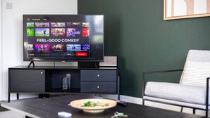A modern living room with a flat-screen TV displaying a streaming service interface, a dark wooden coffee table with a bowl of food, a grey sofa, and a framed picture on the wall. - Elegant 1BR | Balcony | Knightsbridge & Hyde Park (Greater London)