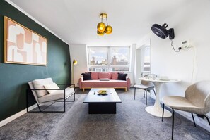 En moderne stue med sofa, lænestole, sofabord og udsigt over byen gennem store vinduer.