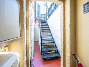 Eine schmale Treppe führt zu einem Raum mit einer weißen Tür, einem roten Boden und einem Metallgeländer.