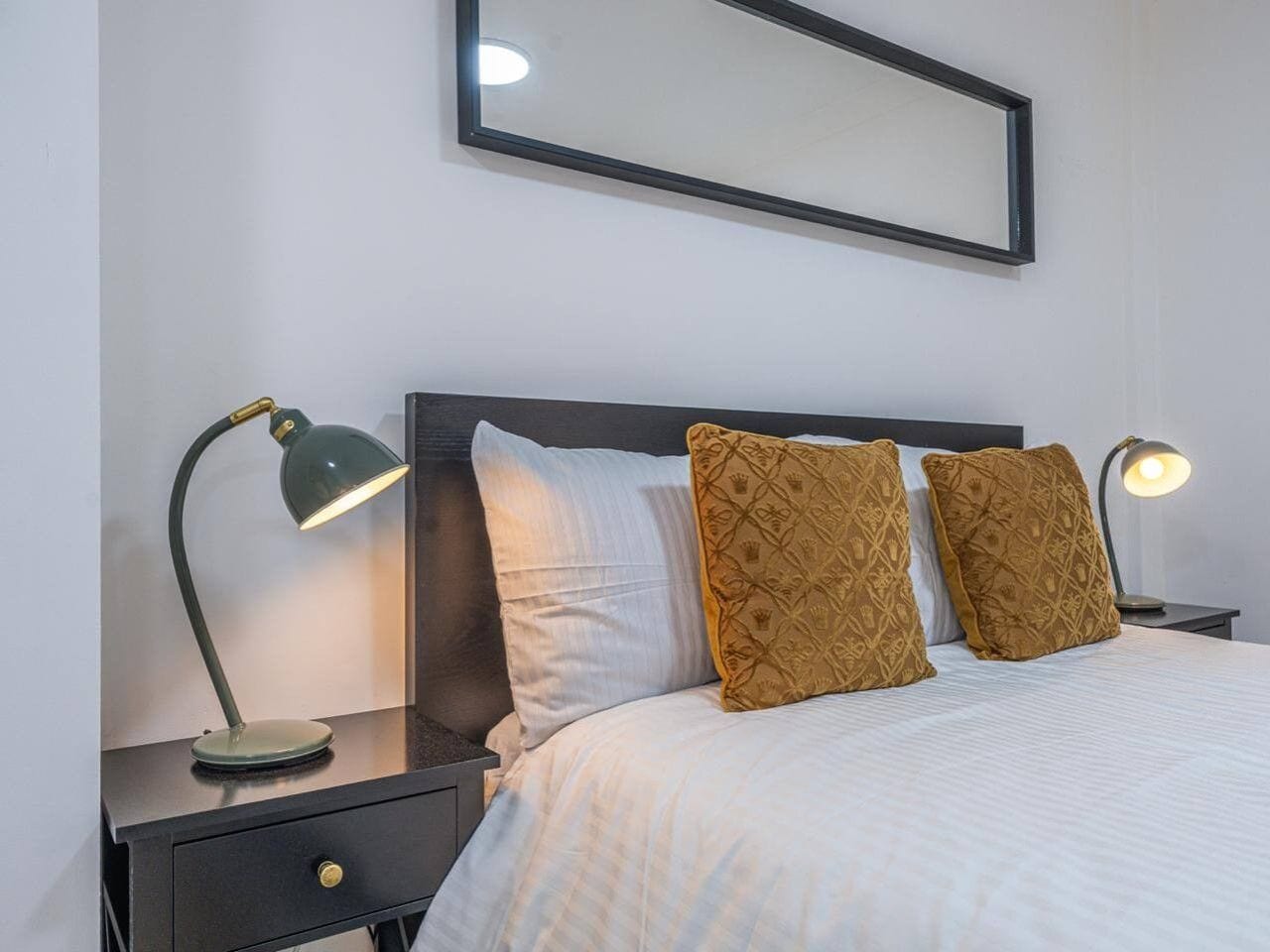A neatly made bed with white bedding and two decorative pillows, flanked by two bedside tables with lamps, one of which is turned on.