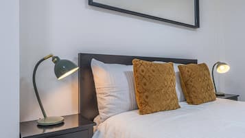 A neatly made bed with white bedding and two decorative pillows, flanked by two bedside tables with lamps, one of which is turned on.