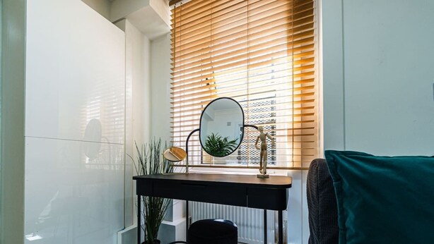 A compact bathroom with a white tiled wall, a black vanity with a mirror, a small stool, and a window with blinds. - Elegant West Kensington Studio | Central London (Greater London)
