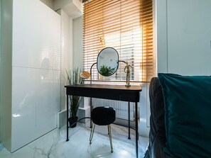Une petite salle de bain avec un mur recouvert de carreaux blancs, un meuble-lavabo noir avec un miroir, un petit tabouret et une fenêtre avec des stores.