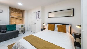 A bedroom with a bed, a sofa, a bedside table, a lamp, a mirror, and a window with blinds. - Elegant West Kensington Studio | Central London (Greater London)