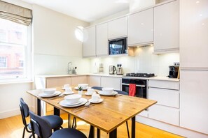 A modern kitchen with white cabinetry, a wooden dining table, and a built-in oven. - Fitzrovia Apartments | 3BR | Sleeps 12 | Oxford-3 (Greater London)