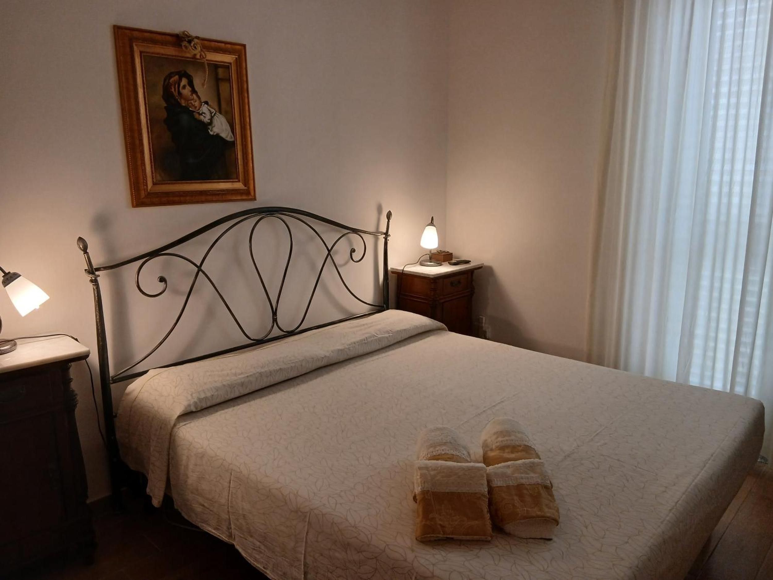Une chambre à coucher avec un lit, une table de chevet, une lampe, un tableau encadré au mur et une fenêtre avec des rideaux.
