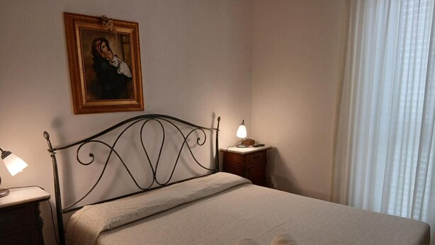 Ein Schlafzimmer mit einem Bett, einem Nachttisch, einer Lampe, einem gerahmten Bild an der Wand und einem Fenster mit Vorhängen.
