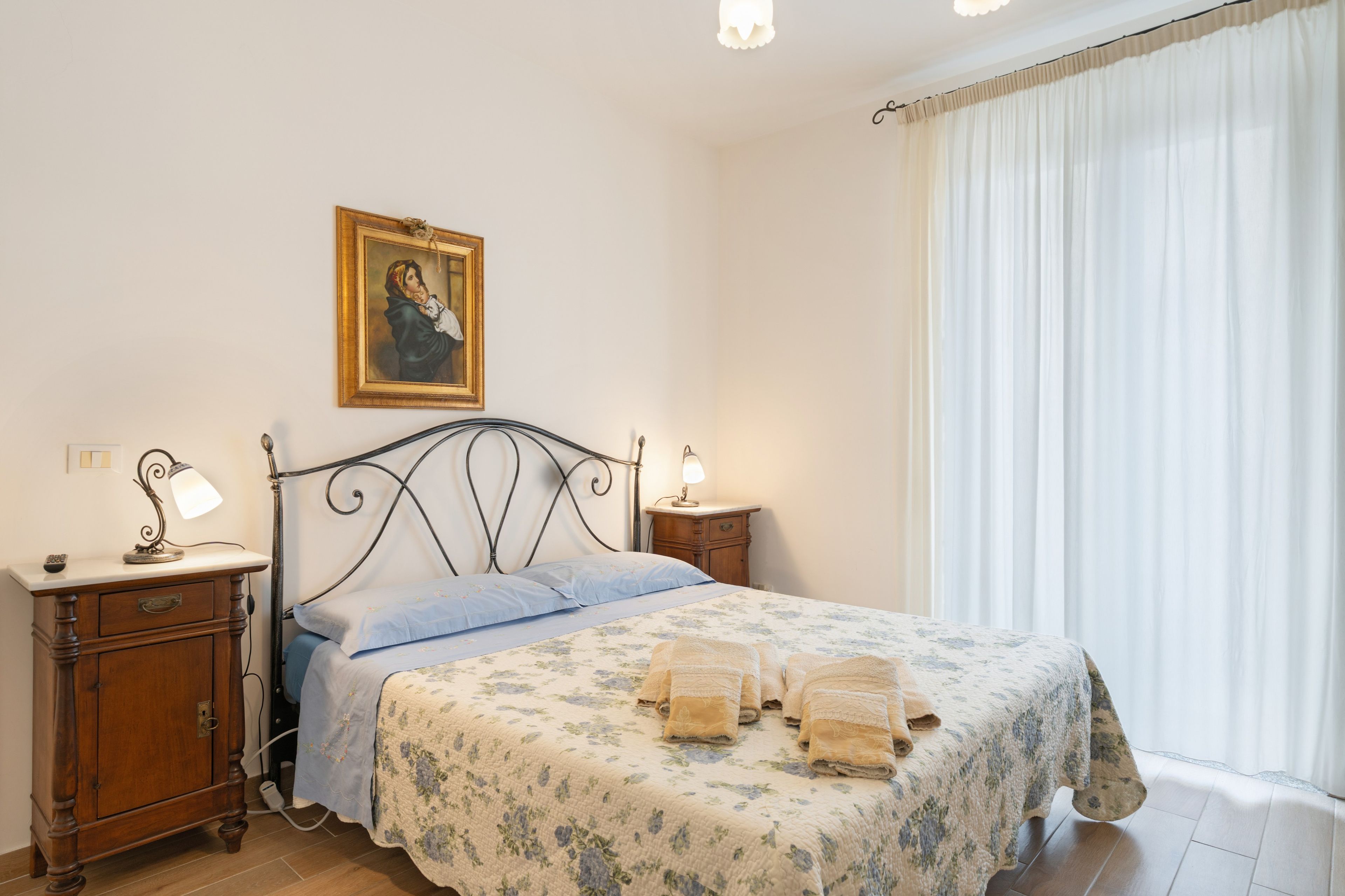 Un dormitorio con una cama, dos mesitas de noche, una lámpara, un cuadro y una ventana con cortinas.