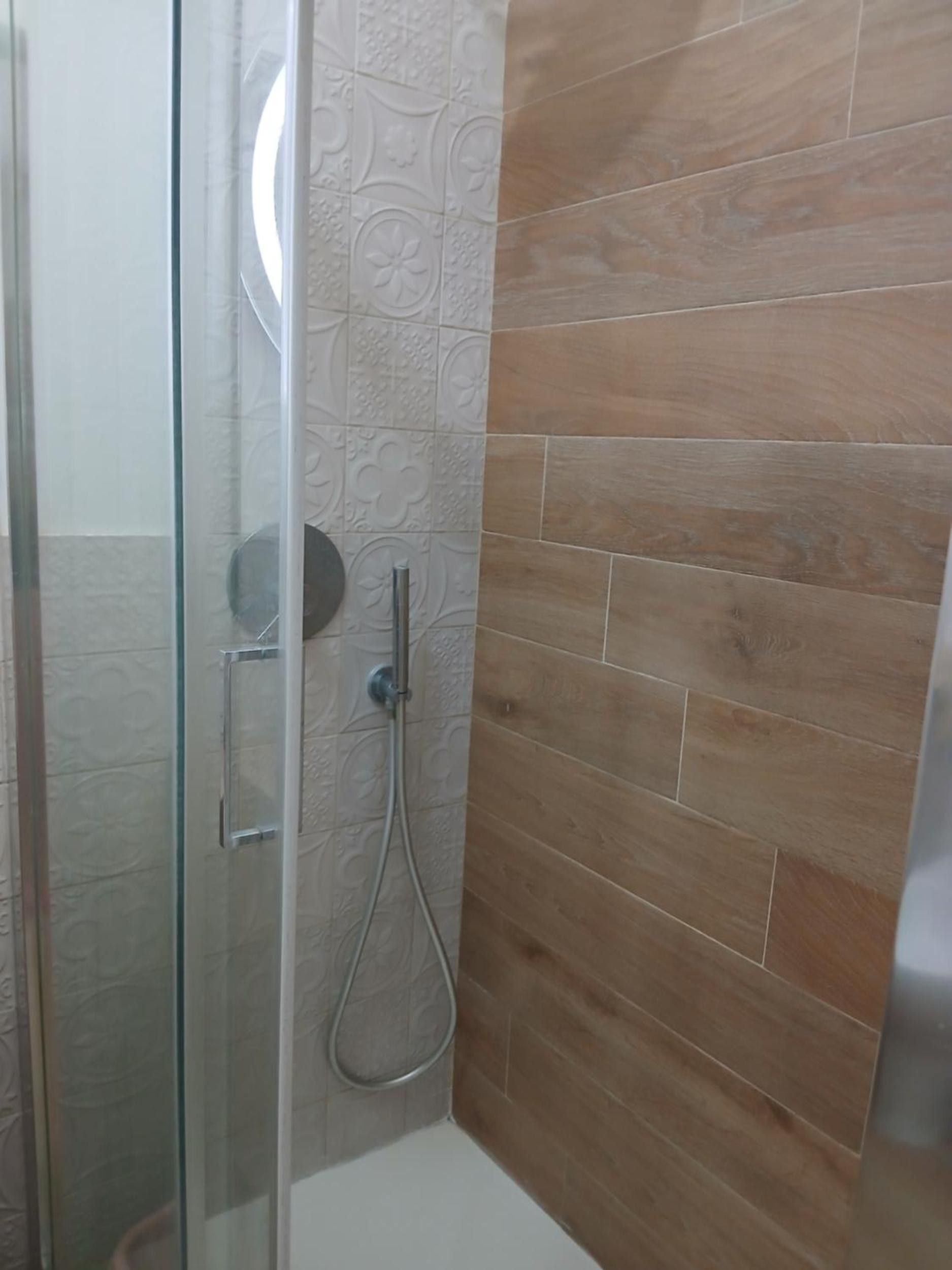 Un espace douche doté d’une porte en verre, d’un pommeau de douche à main et d’un mur recouvert de carreaux décoratifs.