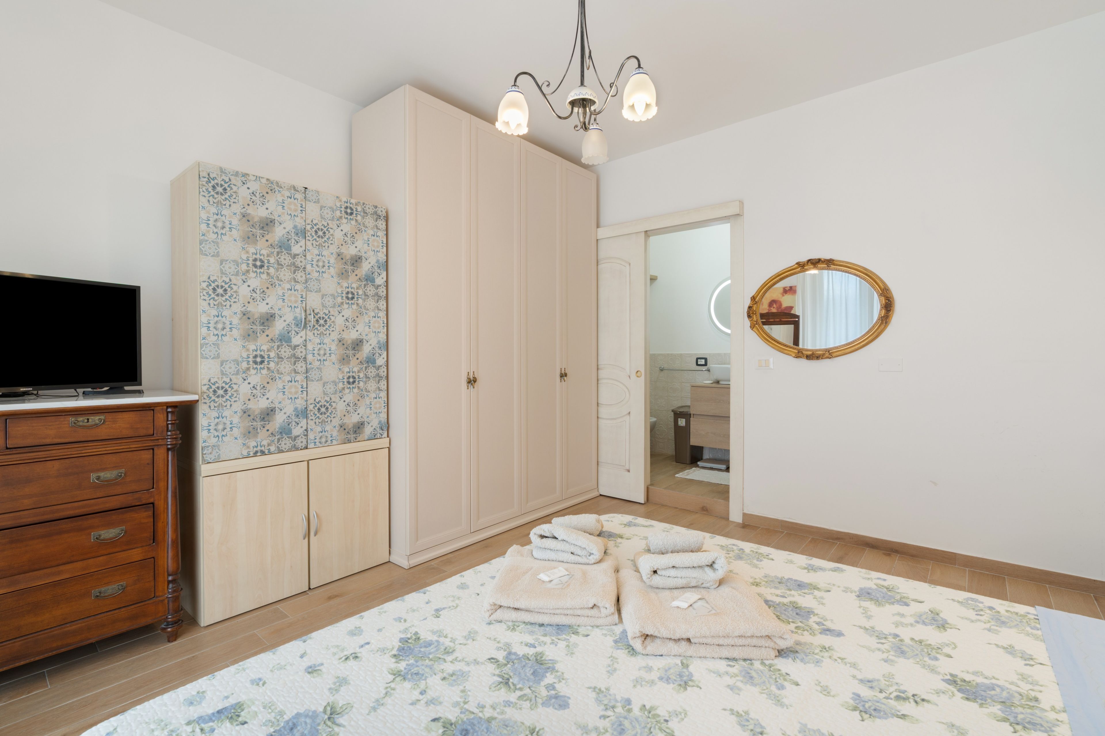 Una camera da letto con una televisione su una cassettiera in legno, un armadio con ante decorate, un lampadario, uno specchio e un bagno visibile attraverso una porta aperta.