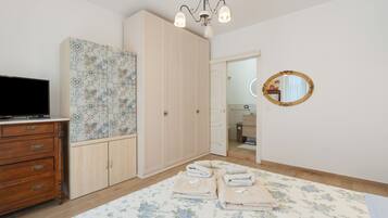 Una camera da letto con una televisione su una cassettiera in legno, un armadio con ante decorate, un lampadario, uno specchio e un bagno visibile attraverso una porta aperta.