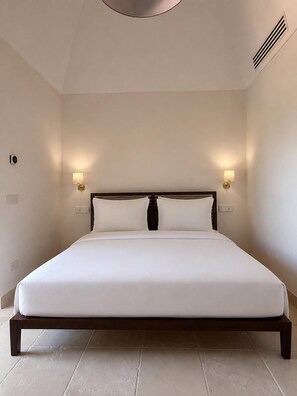 Una cama grande con estructura de madera y ropa de cama blanca, dos lámparas fijadas a la pared y una rejilla de ventilación en el techo.