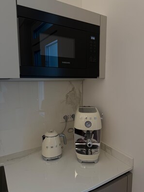 Una cocina con un microondas negro sobre una encimera blanca, una tetera eléctrica blanca y una cafetera blanca.