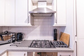 A modern kitchen with white cabinets, a stainless steel range hood, a gas stove, a coffee maker, and a knife block. - Historic City of London Gem | 1 Min to Cannon St (Greater London)