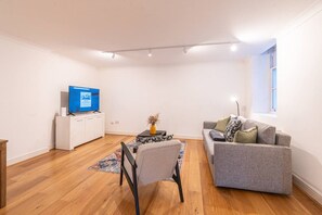Un salon moderne avec un téléviseur à écran plat, un canapé gris, une petite table sur laquelle est posé un vase, et un plancher en bois.