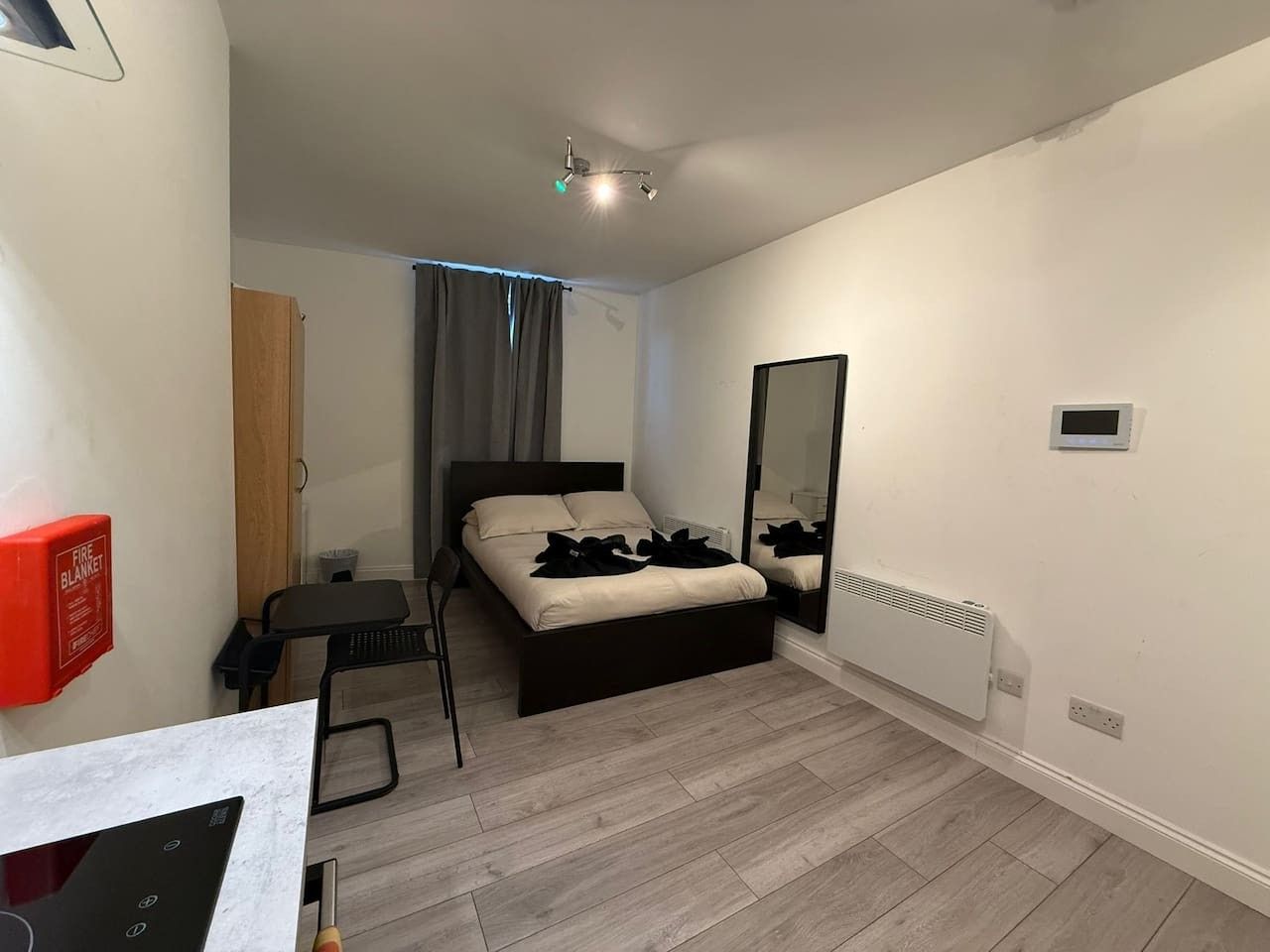 A bedroom with a bed, a desk, a chair, a mirror, a fire blanket, and a radiator.