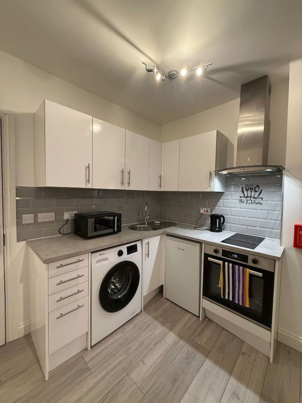 A compact kitchen with white cabinets, a built-in washing machine, a microwave, a sink, and a small oven.